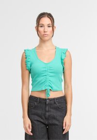 Turkusowy top typu cropped z falbaniastymi rękawami, z marszczonym przodem i zapięciem na sznurek. W połączeniu z ciemnoszarymi, wysokimi spodniami dżinsowymi.