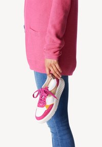 Baskets blanches avec des accents en suède rose et jaune, semelle texturée et lacets roses. Tenues dans une main à côté d'un pull rose et d'un jean en denim.