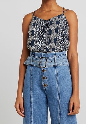 Kobieta nosząca granatowy, wzorzysty top bez rękawów, wpuszczony w niebieskie dżinsy z wysokim stanem, szerokim paskiem i guzikami z przodu.