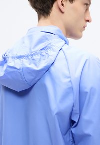 Νεαρός άνδρας που φοράει ανοιχτό μπλε μπουφάν με κουκούλα, εμφανιζόμενος από πίσω και πλάγια μπροστά σε απλό λευκό φόντο.