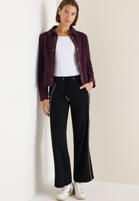 Bordeaux geklede jas met drukknoopjes, witte shirt, zwarte broek met witte zijstripes en witte sneakers met rode accenten.