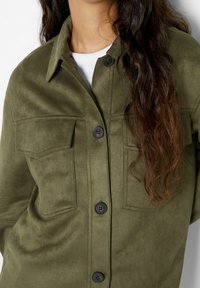 Giacca a camicia verde oliva in morbida pelle scamosciata, con due tasche a pattina e bottoni neri sul davanti.