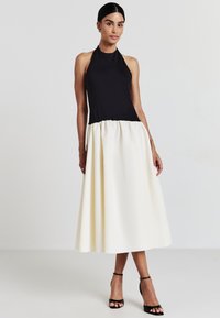 Top a halter nera con una gonna fluida color crema. Il tessuto è liscio con vita arricciata. La modella indossa tacchi neri con cinghie.