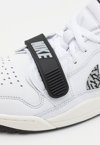 Baskets blanches Nike avec perforations sur l'orteil, lacets blancs, bande noire avec le texte embossé "NIKE", et accent gris à motif près du talon.
