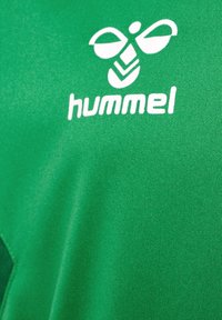 Grön sporttröja i texturerat tyg med en vit "hummel"-logotyp och en bi-liknande emblem ovanför.
