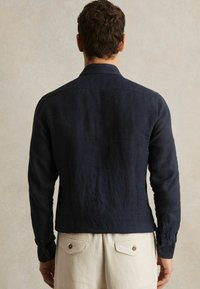 Hombre con cabello oscuro de espaldas con camisa azul marino de manga larga y pantalones beige claro con bolsillos traseros abotonados.
