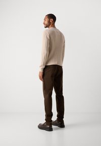 Man stands facing right wearing a beige cable-knit sweater, dark brown pants, and dark brown leather shoes against a plain white background.