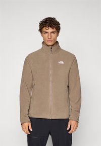 Beige fleece jacket with a high collar, zip front, and two side pockets. Features a white logo on the upper left side. Smooth texture.