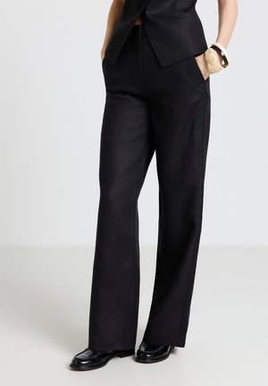 Frau trägt schwarze, weit geschnittene Hose, schwarze glänzende Loafers, schwarzen ärmellosen Blazer und klobige Armbänder am rechten Handgelenk. Hände in den Taschen.