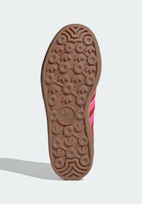 Ruskea kuminen urheilukengän pohja, jossa on texturoitu, kaareva kuviointi, joka sisältää pyörö- ja kolmiomaisia elementtejä, keskellä kohokuvioitu logo.