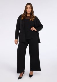 Completo giacca nera con finitura testurizzata, abbinato a pantaloni a gamba larga. Un top trasparente completa l'outfit, accentuato da tacchi neri.