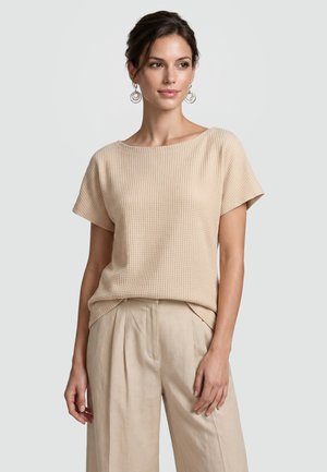 Frau trägt beige strukturiertes Kurzarm-Top und weite Hose, mit runden Anhänger-Ohrringen, vor einem einfachen hellen Hintergrund stehend.