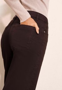 Tamno smeđi traperice s oblikovanim krojem, s stražnjim džepovima i diskretnim detaljima šivanja, prikazane s rukom u džepu.