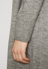 Šedý pletený kousek oděvu s ribbovaným manžetovým zakončením a jemnou strukturou. Ruka se opírá o látku, což zdůrazňuje její tkaní.