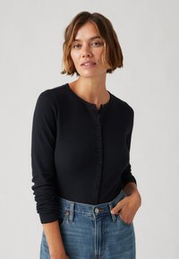 Sieviete ar īsiem brūniem matiem, tērpusies melnā pogājama krekla ar garām piedurknēm un zilos džinsos, pozē ar vienu roku kabatā uz viendabīga fona.