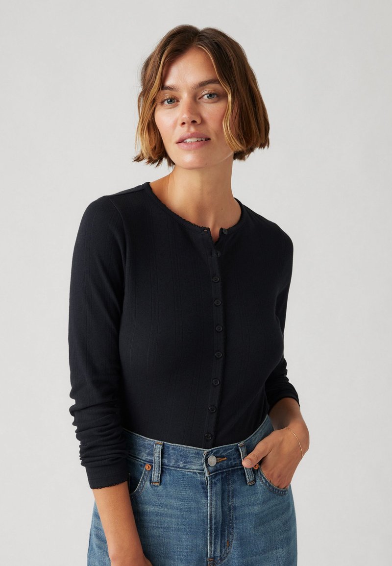 Femme aux cheveux bruns courts portant un haut noir à manches longues boutonné et un jean bleu, posant avec une main dans la poche devant un fond uni.