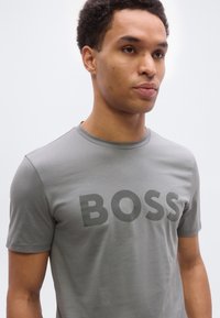 Άνδρας που φοράει ανοιχτό γκρι μπλουζάκι με μεγάλο σκούρο γκρι κείμενο "BOSS", κοιτάζοντας ελαφρώς προς τα δεξιά με φόντο ένα απλό λευκό.