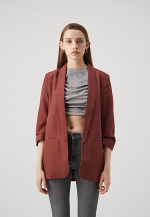 Rostfarbener Blazer mit Schalkragen, offener Front, Dreiviertelärmeln und Fronttaschen, kombiniert mit einem grauen gerippten Crop-Top und grauen Jeans.