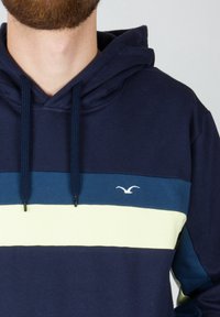 Uomo che indossa un hoodie blu navy con strisce orizzontali blu scuro e giallo chiaro e un piccolo logo di uccello bianco sul petto.