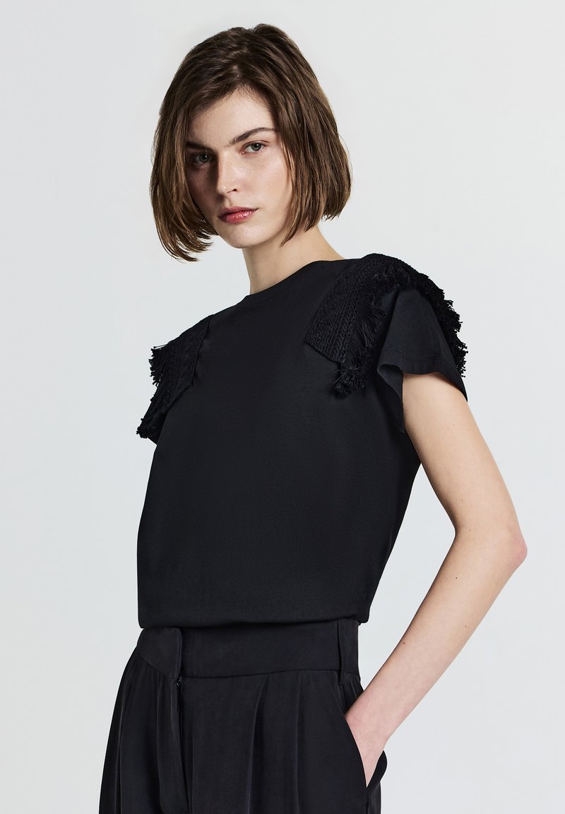 Top nero a maniche corte con accenti frangiati testurizzati sulle spalle, caratterizzato da una vestibilità rilassata e una trama di tessuto liscia. Abbinato a pantaloni a vita alta.