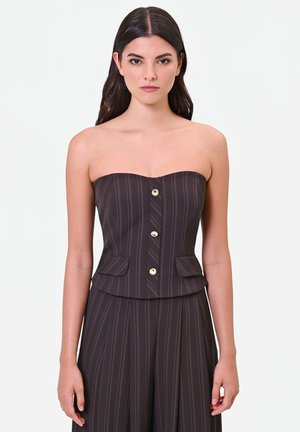 Donna con lunghi capelli castani indossa un top scuro senza spalline a righe sottili con bottoni dorati e pantaloni a vita alta coordinati a righe sottili su sfondo bianco.