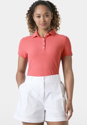 Mujer con cabello rizado que lleva una camiseta polo de manga corta color coral metida en unos pantalones cortos blancos de talle alto, de pie con las manos en los bolsillos.