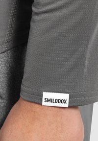 Graues sportliches T-Shirt mit strukturiertem Stoff, versehen mit einem schwarz-weißen Markenetikett mit der Aufschrift "SMILODOX" am Saum.