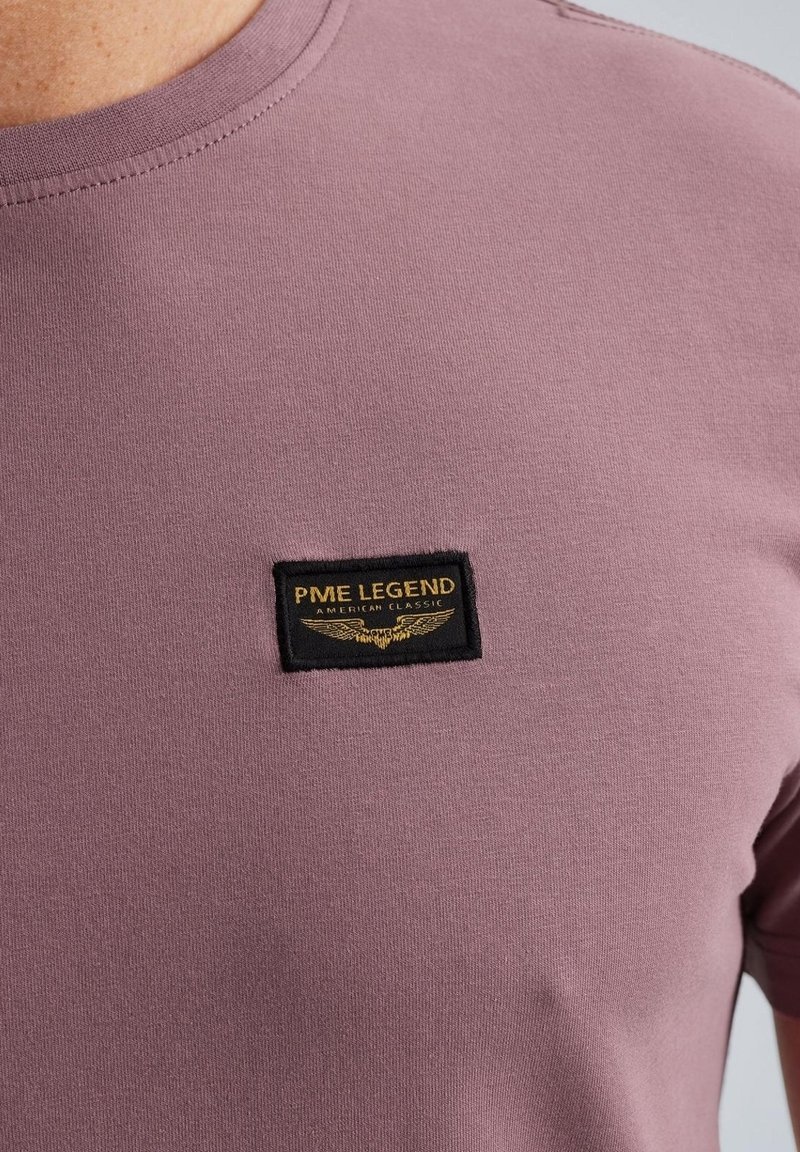 Nærbillede af en mauve t-shirt med et sort og guld PME Legend American Classic patch på brystet nær kraven.