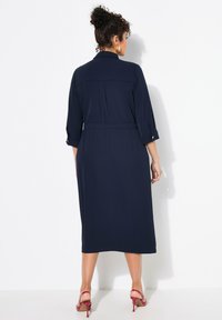 Marineblaues langes Kleid mit Dreiviertelärmeln, tailliertem Schnitt und gefaltetem Rücken. Glattes Material, minimale Hardware-Akzente und midi-lange.