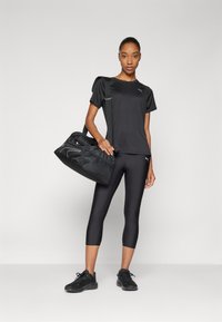 Melna sporta T-kreklā, melnos capri legingos un melnās sneakers. Modelis tur melnu sporta somu ar acu audumu un rāvējslēdzēja nodalījumiem.