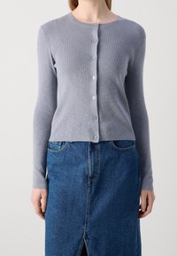 Světlý šedý žebrovaný kardigan s kulatým výstřihem, dlouhými rukávy a zapínáním na knoflíky vpředu, v kombinaci s modrou džínovou sukní s rozparkem.