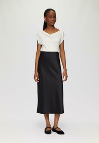 Schwarzer Satin-Midirock mit figurschmeichelndemTaillenbund und gewelltem Saum. Weißes, drapiertes Oberteil mit kurzen Ärmeln. Model trägt schwarze Ballerinas.