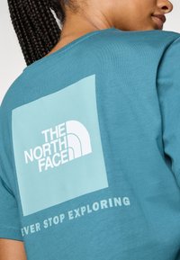 Tirkizna pamučna majica s velikim svijetloplavim kvadratom i bijelim tekstom "THE NORTH FACE" i "EVER STOP EXPLORING" na leđima.