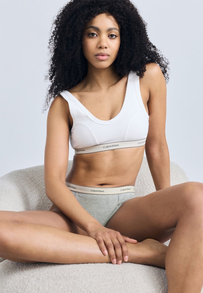 Λευκό βαμβακερό bralette με καμπυλωτή γραμμή λαιμού και λωρίδα με το λογότυπο Calvin Klein. Γκρι βαμβακερά σλιπ με λωρίδα λογότυπου. Μικρή υφή. Το μοντέλο καθισμένο.