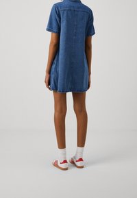 Denim-mekko, jossa on lyhyet hihat, sininen väri, rento malli, takana jalkotuki. Yhdistetty valkoisiin tennareihin, joissa on punaisia yksityiskohtia.