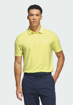Hombre con una camiseta polo de manga corta de color amarillo brillante metida dentro de pantalones azul oscuro con un cinturón negro, de pie contra un fondo liso.