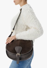 Hnedá kožená crossbody taška s semišovým klopou, zlatými kovovými prvkami a textúrovaným povrchom. Nosená cez biely pletený sveter a džínsy.