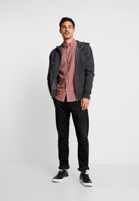 Graue Zip-Hoodie über einem hellrosa Hemd mit Knopfleiste, kombiniert mit schwarzen Jeans und schwarzen Sneakers. Einfaches Design mit neutralen Farben.