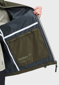 Χέρι κρατάει ανοιχτό ένα χακί πράσινο μπουφάν που δείχνει ένα σκούρο εσωτερικό με μια μεγάλη τσέπη με φερμουάρ με την ετικέτα «RainPack» και λευκές αντανακλαστικές λωρίδες.