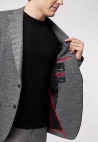 Uomo che indossa un blazer a quadri grigi sopra un maglione nero, mentre tiene aperto il blazer per mostrare la tasca interna bordata di rosso con le etichette del marchio.