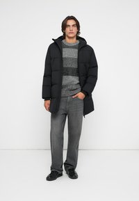 Giacca a vento nera con cappuccio, caratterizzata da un design trapuntato. Maglione grigio a righe sotto, abbinato a jeans grigi e scarpe nere.