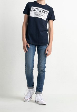Laivansinis t-paita, jossa on valkoinen "PETROL & CO" -grafiikka, yhdistettynä slim fit -sinisiin farkkuihin ja valkoisiin tennareihin. Rentoa, yksinkertaista muotoilua, joka sopii arkeen.