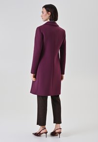 Cappotto di lana borgogna con una vestibilità su misura, maniche lunghe e colletto classico; abbinato a pantaloni marroni a motivo e scarpe con tacco.