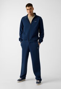 Ensemble de survêtement bleu marine en tissu doux ; comprend une veste zippée et un pantalon à taille élastique. Petit logo "GUESS" sur les deux pièces.