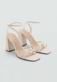 Beige Blockabsatz-Sandalen mit mehreren dünnen Riemen, die vorne überkreuzt sind. Mit einem Knöchelriemen und einer goldenen Schnalle. Glatte Textur.