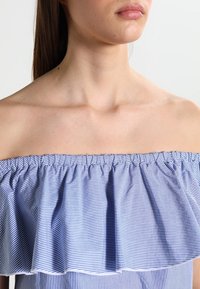 Niebiesko-biały pasek, bluzka bez ramiączek z marszczonym dekoltem i falbaną, wykonana z lekkiego materiału o miękkiej teksturze.