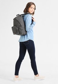 Mochila de tela gris con forma redondeada y bolsillo frontal, llevada por una persona con una camisa de mezclilla azul clara y jeans oscuros.