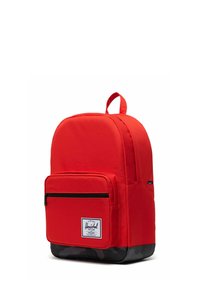 Sac à dos en tissu rouge de forme arrondie, avec une poche avant zippée et un fond noir. Il comporte une poignée supérieure et une étiquette logo à l'avant.