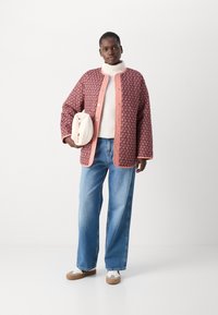Röd mönstrad jacka med rosa detaljer, vit höghalsad tröja, blå vida jeans och beige sneakers. Håller en krämfärgad väska.