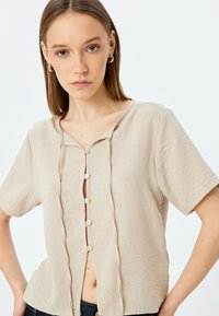 Blusa beige de mangas cortas con escote redondo, cierre frontal de botones, tejido texturizado y pliegues sutiles para dar estructura.